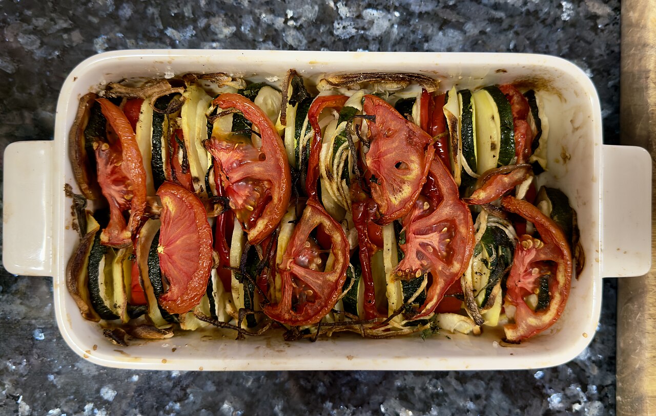 Layered baked potatoes, courgettes, red peppers, tomatoes and onions in a rectangular white baking dish