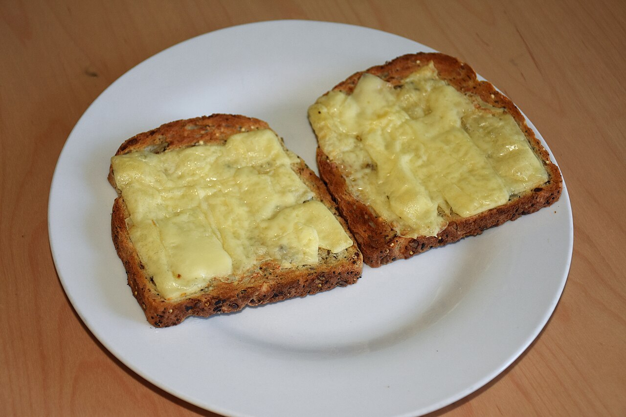 Zwei offene Scheiben Körnerbrot mit geschmolzenem orangefarbenem Cheddar auf einem weißen Teller
